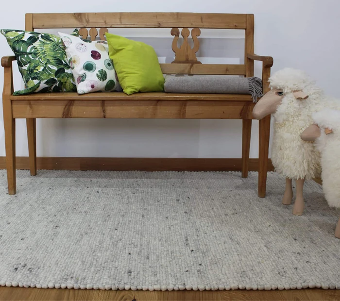 Auf einer Sitzbank liegen 3 Kissen und eine Decke, davor liegt ein Teppich und zwei Deko-Schafe stehen neben der Bank. Produkte der Chiemgauer Weberei.