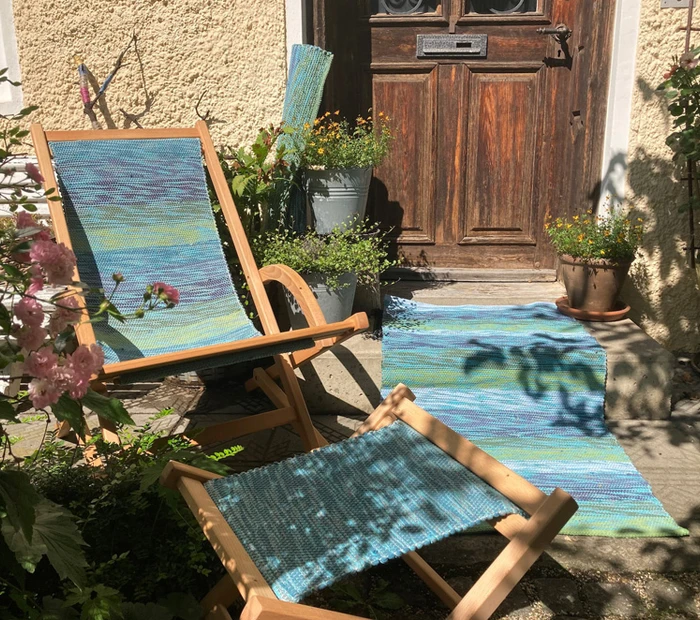 In einem Garten ist ein Teppich, ein Liegestuhl, ein Sitzhocker ausgestellt. Die Bezüge sind von der Chiemgauer Weberei 