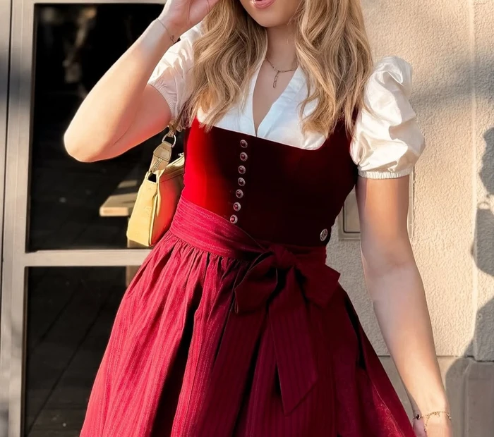 Abgebildet ist ein rotes Samtdirndl mit weißer Dirndlbluse, erhältlich in der Chiceria Prien, solange Vorrat reicht.